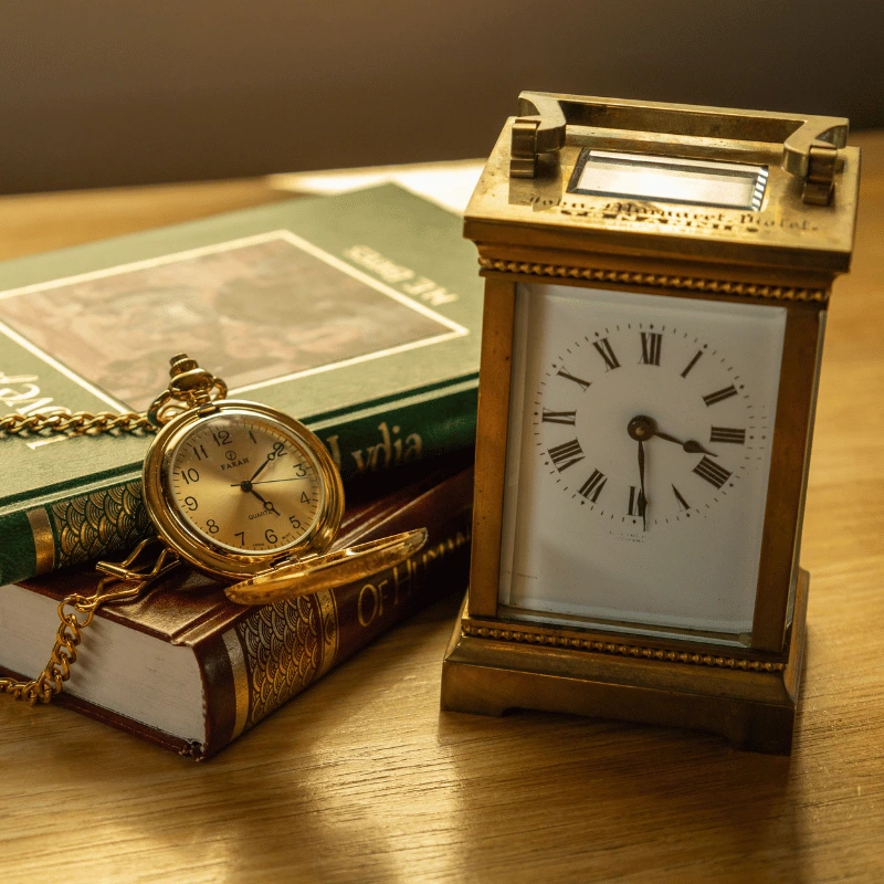 Rectangular antique gold clock and gold pocket watch displayed alongside two historical books, showcasing a blend of timeless craftsmanship and rich history.